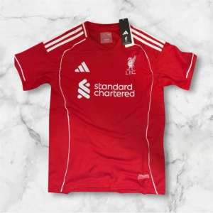 Liverpool röd fotbollströja Adidas - Säljer en röd Liverpool fotbollströja från Adidas med vita detaljer och logga på bröstet. Tröjan har korta ärmar, tre vita ränder på axlarna och Standard Chartered tryckt på framsidan. Perfekt för dig som älskar fotboll och vill visa ditt stöd för LFC.
