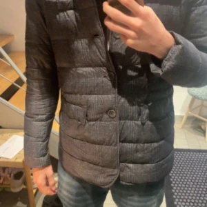 Grå rutig dunjacka från Herno - Snygg grå dunjacka från Herno med diskret rutigt mönster. Jackan har klassisk krage, knäppning med tre knappar och två stora fickor framtill. Den är quiltad och har en stilren look som passar perfekt till jeans. Perfekt för kyliga dagar.