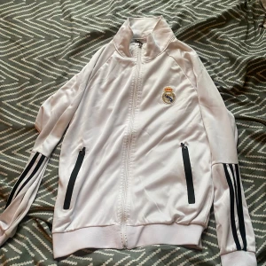 Vit Real Madrid ziptröja Adidas - Säljer en vit ziptröja från Adidas med Real Madrid-logga på bröstet. Tröjan har svarta ränder längs ärmarna, två fickor med dragkedja och hög krage. Perfekt för dig som älskar fotboll och vill ha en sportig look.