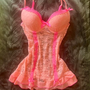 Rosa spetstopp med rosetter - Säljer en supersöt rosa topp med spets och flera små rosetter. Toppen har axelband och är dekorerad med ljusrosa och starkrosa detaljer, samt en volangkant längst ner. Perfekt för dig som gillar romantisk och feminin stil.