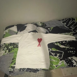 Vit stickad tröja från Ami Paris - Säljer en vit stickad tröja från Ami Paris med röd hjärt- och A-detalj på bröstet. Tröjan har rund halsringning och långa ärmar, perfekt för dig som gillar stilrena plagg med en twist. Passar lika bra till jeans som till kjol.