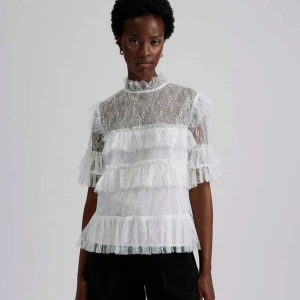 Vit spetsblus med volanger - Nypris 1800kr. Supersöt vit blus i spets med flera lager volanger och hög hals från By Malina. Blusen har korta ärmar med volangdetaljer och är genombruten i materialet för en romantisk vibe. Perfekt för dig som gillar detaljer och vill sticka ut lite extra. Köp på plick aldrig använd. 