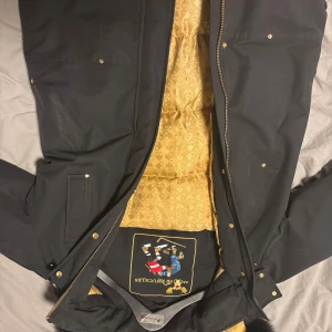 Svart pufferjacka från Moose Knuckles - Säljer en svart pufferjacka från Moose Knuckles med guldfärgat, mönstrat foder och snygga gulddetaljer. Jackan har dragkedja, knappar och ikonisk logga på ärmen. Perfekt för dig som vill ha en varm och stilren jacka med exklusiv känsla.