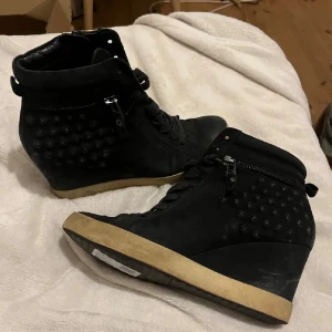 Svarta kilklackade boots med nitar - Säljer ett par svarta boots med kilklack och coola nitar på sidorna. Skorna har snörning framtill och dragkedja på sidan för enkel på- och avtagning. Yttermaterialet är mocka och sulan är beige. Perfekt för dig som gillar edgy stil. Sulan är borttagen men man kan ha utan 