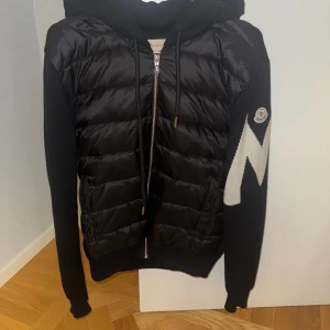 Svart Moncler cardigan - Svart pufferjacka från Moncler med stickade ärmar och stor vit M-detalj på ena armen. Jackan har huva, dragkedja framtill och Moncler-logga på ärmen. Materialet är en mix av dun och stickad bomull, perfekt för dig som vill ha en snygg jacka nu under hösten