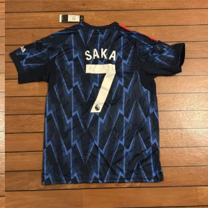 Arsenal Saka #7 blå Adidas fotbollströja - Snygg Arsenal fotbollströja från Adidas i slim fit, med Saka och nummer 7 på ryggen. Tröjan är mörkblå med ett elektriskt zigzag-mönster i blått, röda ränder på axlarna och vita tryck. Tillverkad i lätt polyester, perfekt för match eller träning.
