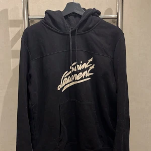 Svart Saint Laurent hoodie - Svart hoodie från Saint Laurent i storlek S med vit logotyp på bröstet. Tröjan har huva med snörning, skriv vid frågor!