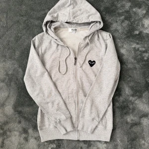 Grå hoodie Comme des Garcons Play - Grå zip-hoodie från Comme des Garçons Play med svart hjärta på bröstet och stort tryck på ryggen. Storlek S, passar som XS – unik modell som man sällan ser.