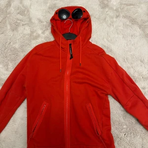 Röd hoodiejacka från C.P. Company - Säljer en röd hoodiejacka från C.P. Company med ikoniska goggles på huvan. Jackan har dragkedja framtill, två fickor med dragkedja och snörning i huvan. Materialet är mjuk bomull och passformen är normal. Perfekt för dig som vill sticka ut med streetwear-vibe.