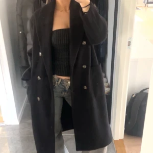 Svart lång kappa med knappar - Säljer en svart lång kappa med dubbelknäppning och bred krage. Jackan har stora fickor på sidorna och en stilren, rak passform som ger en chill vibe. Perfekt för dig som gillar minimalistisk stil och vill ha något som funkar hela hösten och vintern.