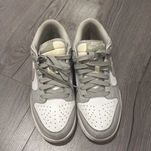 Nike Dunk Low i grått och vitt - Nike Dunk Low sneakers i stilren grå och vit färgkombination. Skorna har klassisk snörning, perforerad tå och ikonisk Swoosh på sidorna. Ovandel i skinn och syntet, låg profil och bekväm platt sula. Perfekta för dig som gillar clean och tidlös streetstyle.