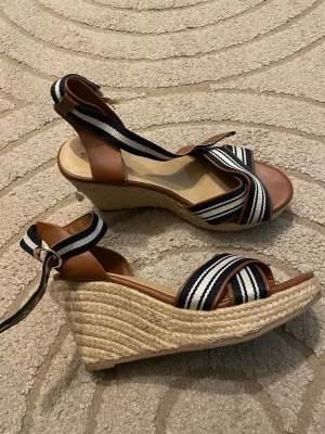 Randiga kilklackade sandaler - Snygga sandaler med kilklack och remmar i brunt skinn och randigt tyg i svart, vitt och blått. Remmarna går över foten och runt ankeln med spänne. Sulan är täckt med flätad jute för en somrig känsla. Använd gärna köp nu❤️