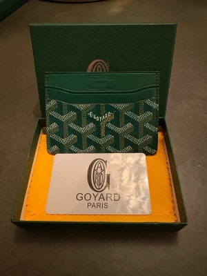 Grön korthållare från G - Snygg grön korthållare från G med ikoniskt mönster och flera kortfack. Tillverkad i exklusivt skinn med Goyards klassiska logga framtill. Perfekt för dig som vill ha en stilren och lyxig accessoar i fickformat.