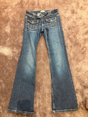 Blå bootcut jeans med fickdetaljer - Snygga blå bootcut jeans med dubbla knappar och framfickor med lock och knappar. Jeansen har markerade sömmar och klassisk femficksdesign. Perfekta för dig som gillar en retroinspirerad look. Storlek 152cm