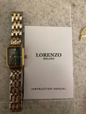 Lorenzo Milano klocka guld/silver - Stilren klocka från Lorenzo Milano med rektangulärt svart ur, guldfärgad boett och tvåfärgad länk i guld och silver. Klockan har en klassisk design med metallarmband och minimalistisk urtavla. Perfekt accessoar för dig som gillar en elegant look. Du får den med boxen och allt