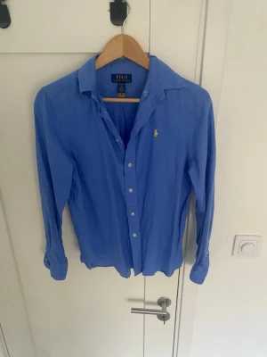 Blå linneskjorta från Polo Ralph Lauren. - Snygg blå skjorta från Polo Ralph Lauren i linne med klassisk krage och knappar framtill. Ikoniska gula loggan broderad på bröstet. Perfekt för dig som gillar en avslappnad men ändå stilren look. Skjortan är i L i barn (160)