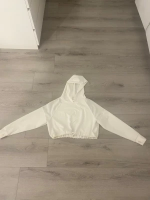 Vit croppad hoodie - Säljer en vit croppad hoodie med långa ärmar och elastisk midja. Hoodien har en enkel och clean design utan synligt märke eller tryck. Perfekt för en chill och avslappnad stil. Materialet ser ut att vara mjukt och bekvämt, troligen bomullsblandning.