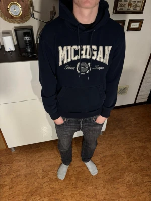 Mörkblå Michigan hoodie med tryck - Snygg mörkblå hoodie från Jack and Jones  med stor Michigan-text i vitt framtill och detaljerad logga under. Klassisk känguruficka och justerbar huva med snören. Tillverkad i mjukt bomullsmaterial, perfekt för en avslappnad streetwear-look. J
