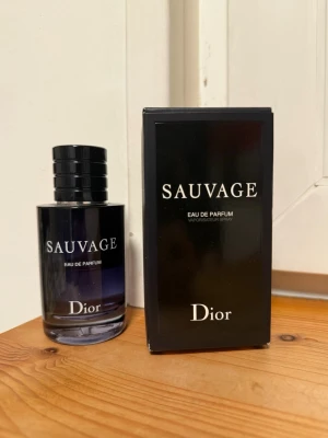 Dior Sauvage Eau de Parfum - Dior Sauvage Eau de Parfum med elegant svart och mörkblå glasflaska och matchande svart kartong. Flaskan har en stilren, rund form med silverdetaljer på korken och tydlig logga framtill. En ikonisk doft som utstrålar lyx och modernitet. UNGEFÄR HÄLFTEN KVAR 
