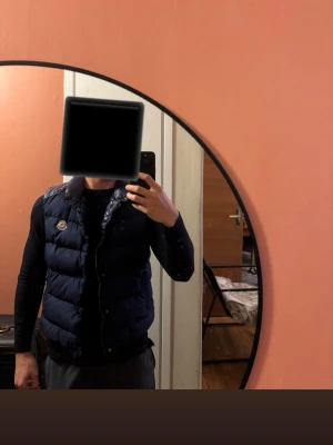 Mörkblå dunväst från Moncler - Snygg mörkblå dunväst från Moncler med hög krage och två dragkedjeförsedda fickor. Västen har ett klassiskt quiltat mönster och Moncler-logga på bröstet. Perfekt att slänga över en hoodie eller tröja för extra värme och stil. Storlek S men passar mer som en xs, buda gärna 