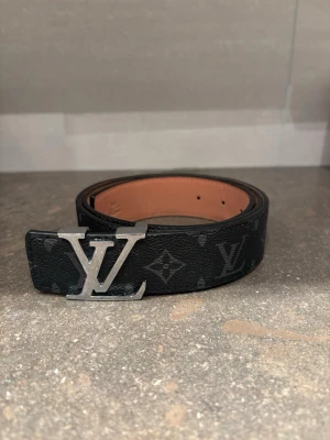  monogram bälte svart silver bulka - Snyggt svart bälte, klassiskt monogrammönster och stor silver spänne. Bältet är gjort i läder med beige insida och har exklusiv känsla. Perfekt accessoar för att lyfta din outfit.