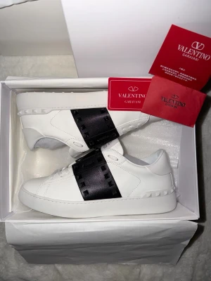 Valentino skor - Snygga vita Valentino Garavani Open sneakers med bred svart rem över vristen och ikoniska nitar på hälen. Skorna har rund tå, platt sula och är tillverkade i skinn. Perfekta för dig som vill ha en clean men ändå lyxig look.