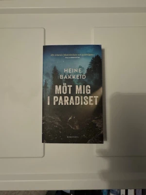 Möt mig i paradiset av Heine Bakkeid - En spännande deckare av Heine Bakkeid med titeln 'Möt mig i paradiset'. Omslaget har en mörk, mystisk skogsmiljö i blå och svarta toner. Boken är i pocketformat och tryckt på papper. Perfekt för dig som gillar nervkittlande berättelser.