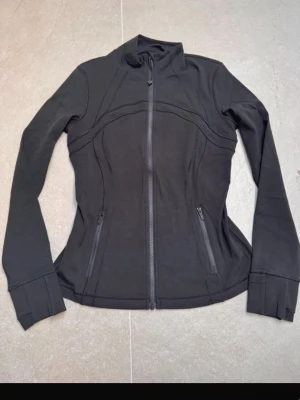 Svart vindjacka från Lululemon - Snygg svart vindjacka från Lululemon med figurnära passform och hel dragkedja framtill. Jackan har långa ärmar, hög krage och två fickor med dragkedja. Perfekt för en aktiv vardag och stilren look. Den är även jätte bra för träning. Det finns även kvitto.