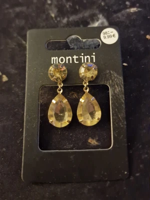 Guldiga droppformade örhängen Montini - Snygga örhängen från Montini med stora, droppformade stenar i guldton. Örhängena har en rund sten upptill och en större, facetterad droppe som hänger ned. Perfekta för dig som vill ha ett statement-smycke med glittrig känsla.