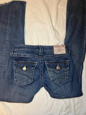 Blå True Religion bootcut jeans - Säljer ett par blå jeans från True Religion med klassiska kontrastsömmar och snygga ficklock bak med silverknappar. Modellen har bootcut och låg midja, tillverkade i bomull med jeansmaterial. Perfekta för dig som gillar detaljer och ikonisk design.