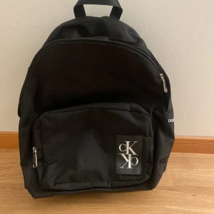 Svart ryggsäck från Calvin Klein - Snygg svart ryggsäck från Calvin Klein med stort CK-logotyp på frontfickan. Ryggsäcken har en klassisk rundad form, dragkedjor i silver och en praktisk ytterficka. Perfekt för skola eller cityhäng. Knappt använd 1-3 gånger 