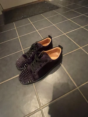 Svarta sneakers med nitar och röd sula - Unika svarta sneakers med coola nitar på tån och sidorna. Skorna har snörning, rund tå och en ikonisk röd sula. Materialet ser ut att vara mocka eller liknande syntet. Perfekta för dig som vill sticka ut med din stil.