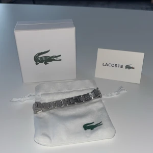 Silverfärgat armband från Lacoste - Snyggt silverfärgat armband från Lacoste i rostfritt stål. Armbandet har en stilren design med länkstruktur och diskret Lacoste-logga. Kommer med original ask och tygpåse med krokodil-loggan. Perfekt accessoar för dig som gillar klassisk och sportig stil.