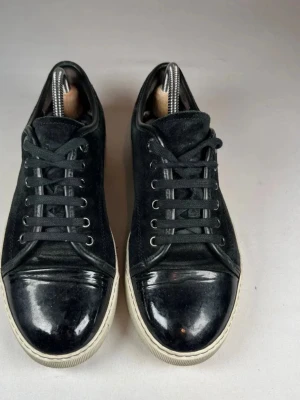 Svarta lanvins  - Stilrena svarta sneakers med ovandel i mocka och blanka detaljer på tån. Skorna har snörning och en vit, platt sula som ger en klassisk look. Perfekta för dig som gillar en enkel men snygg stil.