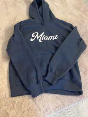 Mörkblå hoodie med Miami-tryck - Snygg mörkblå hoodie från 157 med vit text 'Miami' på bröstet. Klassisk modell med huva och ribbade muddar vid ärmslut och nederkant. Perfekt för chill dagar eller när du vill ha en avslappnad stil.