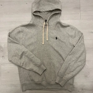 Grå hoodie från Polo Ralph Lauren - Klassisk grå hoodie från Polo Ralph Lauren med broderad logga på bröstet. Hoodien har huva med vita dragsnören, känguruficka framtill och ribbade muddar vid ärmar och nederkant. Tillverkad i mjukt sweatshirtmaterial, perfekt för en avslappnad stil.