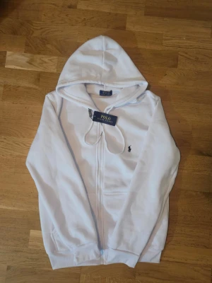 Vit hoodie från Polo Ralph Lauren - Snygg vit hoodie från Polo Ralph Lauren med dragkedja framtill och klassisk huva. Ikoniska broderade loggan i svart på bröstet. Mjuk bomullskänsla och ribbade muddar vid ärmslut och nederkant. Perfekt för en clean och stilren look.