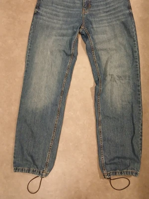 Blå baggy jeans från Ecko Unltd - Säljer ett par blå baggy jeans från Ecko Unltd med broderade detaljer på bakfickorna och justerbara snören vid bensluten. Klassisk femficksmodell i jeansmaterial och avslappnad passform som ger en cool streetwear-look.