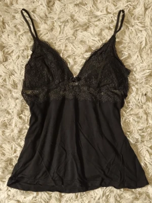 Svart spetslinne med tunna axelband - Svart linne med tunna axelband och v-ringad spets upptill. Överdelen är dekorerad med blommig spets och resten av linnet är i mjukt tyg. Perfekt för lager på lager eller som ett snyggt underklädesplagg.