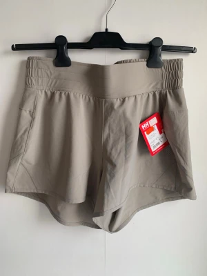 Beige träningsshorts Helly Hansen - Snygga beige träningsshorts från Helly Hansen med bred resår i midjan och praktiska fickor, en sportig look och diskret logga på sidan,  en liten ficka med dragkedja baktill. Tillverkade i ett lätt och stretchigt material.  Samfraktar. 