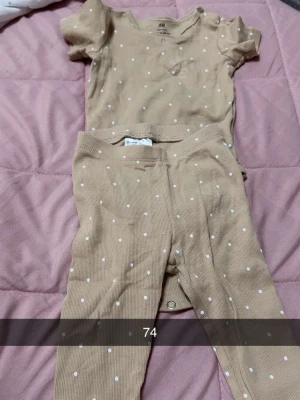 Beige prickigt set från H&M - Sött tvådelat set från H&M i beige med vita prickar. Setet består av en kortärmad body med rund hals och tryckknappar samt matchande byxor i mjukt ribbat material. Perfekt för en bekväm och gullig vardagslook.