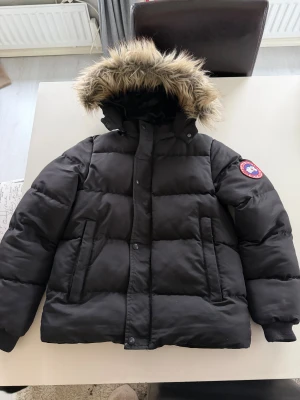 Svart vinterjacka från Canada Goose - Säljer en svart vinterjacka från Canada Goose med ikonisk patch på ärmen. Jackan har quiltad design, dragkedja och knappar framtill samt två snedställda fickor. Perfekt för kalla dagar och riktigt snygg streetstil.