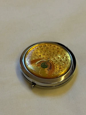 Guldig sminkspegel med påfågelmotiv - Köpt utomlands ett par år sedan - verkligen unik design. Snygg rund sminkspegel i metall med ett detaljerat påfågelmotiv i guld, grönt och koppar på locket. Spegeln är kompakt och har två speglar inuti, perfekt att ha i väskan eller fickan. Elegant design som sticker ut och känns lyxig. Endast defekt är att låset inte funkar. 
