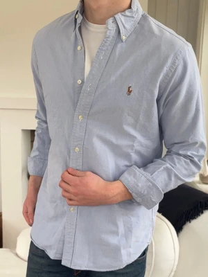 Ljusblå skjorta från Ralph Lauren - Klassisk ljusblå skjorta från Ralph Lauren med button-down krage och broderad logga på bröstet. Skjortan har långa ärmar, vita knappar och är tillverkad i mjuk bomull. Perfekt till jeans eller chinos för en clean och stilren look.