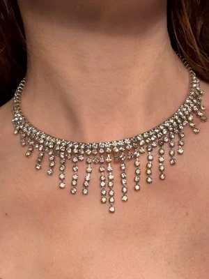 Strasshalsband Med Droppformade Hängen Choker Silver Glitter - Glittrande halsband i silverfärgad metall med flera rader av klara strasstenar och hängande droppformade detaljer. Halsbandet har en choker-modell som sitter nära halsen utan att sitta för tight. Ger en lyxig och trendig känsla. Perfekt för att lyfta vilken outfit som helst. Köpt på Glitter.