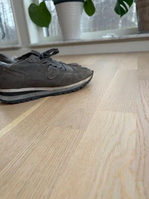 Grå Philippe Model sneakers mocka - Snygga sneakers från Philippe Model i grå mocka med matchande grå skosnören och svartvit sula. Klassisk retroinspirerad design med låg profil och diskret logga på sidan. Perfekta för dig som gillar stilrena och tidlösa sneakers.