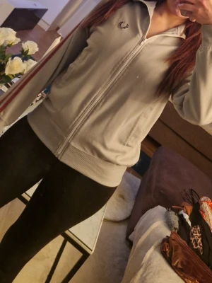 Grå zip hoodie från Fred Perry - Snygg grå zip hoodie från Fred Perry med vit dragkedja och klassisk logga på bröstet. Tröjan har ribbade muddar, huva och röda samt vita ränder längs ärmarna. Perfekt för en sportig och avslappnad stil.