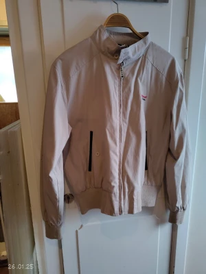 Beige vindjacka med dragkedja - Stilren beige vindjacka från Petroff co.inc med hög krage och dragkedja framtill. Jackan har svarta detaljer vid fickorna och ribbade muddar vid ärmslut och midja. Perfekt för blåsiga dagar och enkel att matcha med olika outfits.