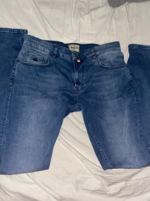 Blåa jeans från Morris - Säljer ett par klassiska blå jeans från Morris med raka ben och snygga kontrastsömmar. Jeansen har fem fickor och en läderpatch bak i midjan. Perfekta för dig som gillar en enkel och tidlös stil.