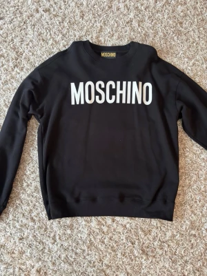 Svart sweatshirt från Moschino - Snygg svart sweatshirt från Moschino med stor vit logga över bröstet. Klassisk rund halsringning och ribbade muddar vid ärmslut och nederkant. Perfekt för en trendig och avslappnad stil.
